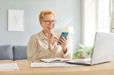 Happy elderly woman working online from home office, laughing, using a phone and internet. Highlighting happiness, productivity, and the role of an accountant in a remote work setting.