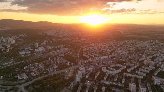 Aerial drone sunset view of sprawling urban residential district nestled between rolling hills with brilliant orange sun low on horizon casting golden light over white apartment blocks and distant mou