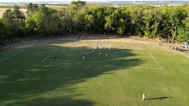 High-angle aerial drone view of a local football match on a green grass field, with players in yellow and white jerseys spreading out in midfield while long tree shadows stretch across the pitch in wa