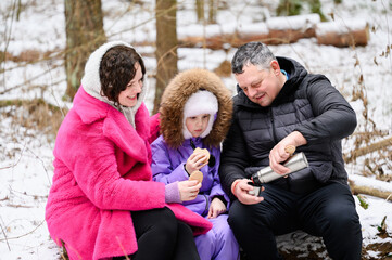 Family with child sits on fallen tree during rest stop, drinking hot tea from thermos with cookies. Parents and girl are having picnic in winter forest. Concept for snacking while hiking.