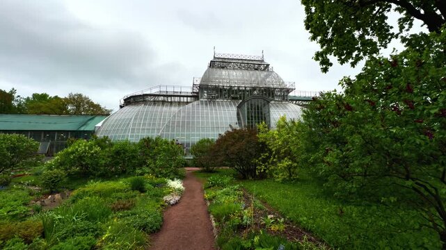 Symmetrical wide shot of the majestic, curved glass facade of the historic greenhouse complex, with a central dirt path leading towards it, surrounded by lush green foliage and flowering plants under