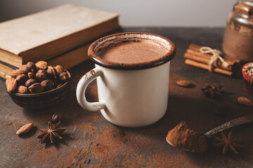 Hot chocolate in white enamel mug on dark rustic surface with nuts, cocoa powder, cinnamon sticks and cupcake, seasonal cozy winter drink concept