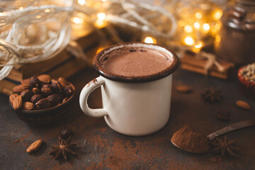 Hot chocolate in white enamel mug on dark surface with nuts, cocoa powder, spices, cupcake and festive lights, seasonal cozy drink and winter holiday concept