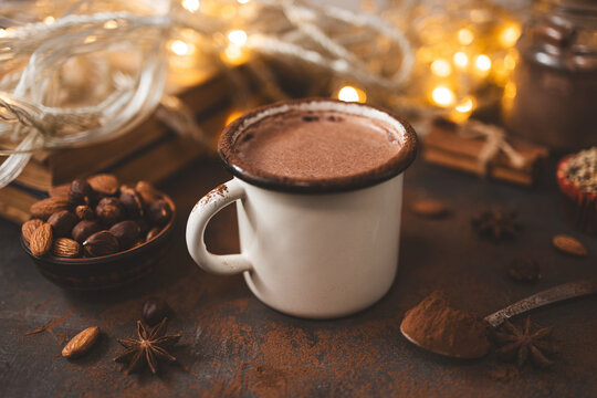 Hot chocolate in white enamel mug on dark surface with nuts, cocoa powder, spices, cupcake and festive lights, seasonal cozy drink and winter holiday concept