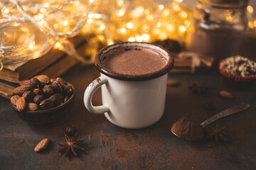 Hot chocolate in white enamel mug on dark surface with nuts, cocoa powder, spices, cupcake and festive lights, seasonal cozy drink and winter holiday concept