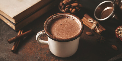 Hot chocolate in white enamel mug on dark rustic surface with nuts, cocoa powder, cinnamon sticks...