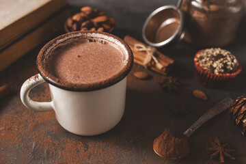 Hot chocolate in white enamel mug on dark rustic surface with nuts, cocoa powder, cinnamon sticks...