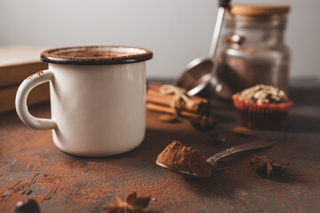 Hot chocolate in white enamel mug on dark rustic surface with nuts, cocoa powder, cinnamon sticks...
