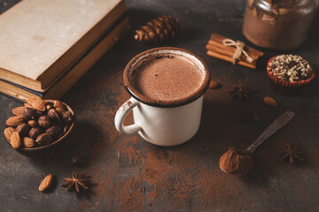 Hot chocolate in white enamel mug on dark rustic surface with nuts, cocoa powder, cinnamon sticks...