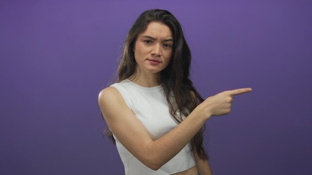 Young hispanic woman with furrowed brow pointing finger toward viewer in purple studio; rebuke anger.