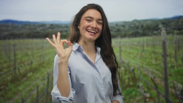 Smiling young hispanic woman makes ok sign with hand among lush green vineyard rows at forest edge; optimism.