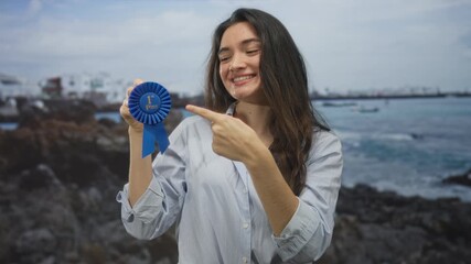 Woman holding first place rosette with finger to forehead l gesture at beach while smiling; ironic triumph.