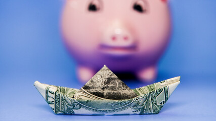 Creative money boat made from a dollar bill in front of a piggy bank on a blue background