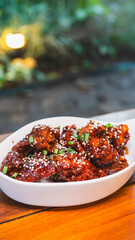 Sizzling, succulent Korean sticky chicken glistening with sauce, topped with sesame seeds and spring onions. A vibrant meal, beautifully presented in an elegant bowl within a stunning ambient setting.