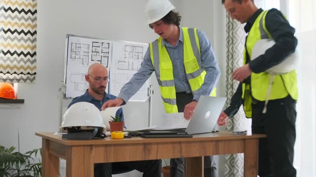 caucasian foreman presenting large blueprint to seated client, two contractors standing, helmet on desk, light filled room, detailed floorplans on wall, attentive client response, collaborative review