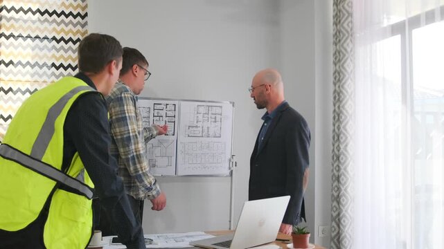 site supervisor joins architects at whiteboard, three professionals study blueprint pinned on wall, collaborative review with plans on table and laptop, focused problem solving and role coordination