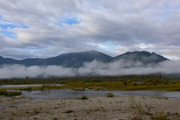 The snow-capped mountains of Altai rise in the background, partially hidden by clouds. In the foreground is a grassy field.