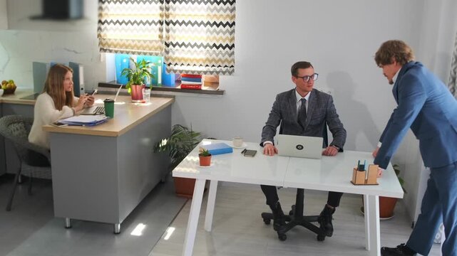negotiation between colleagues in office, business negotiation happening across desk with observing assistant present, businessman and colleague negotiate at desk while assistant observes in office