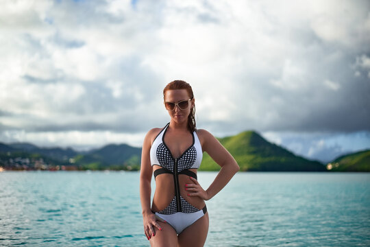 
Attractive woman in a black and white monokini, wearing sunglasses, standing in the ocean with green tropical islands and a cloudy sky in the background - Powered by Adobe