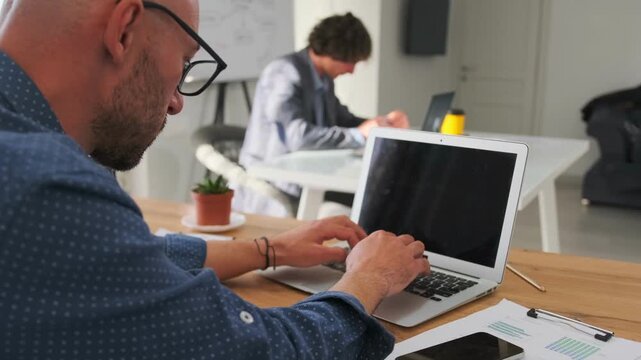 freelancer working alone, individual freelancer intently typing in collaborative office environment, focused white male freelancer with bald head works quietly in shared office space with colleagues