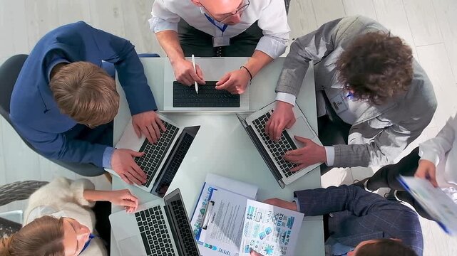 team collaborating over colorful charts, energetic group working together at laptopfilled table, collaborative team engaged in lively brainstorming around triangular table with laptops and colorful