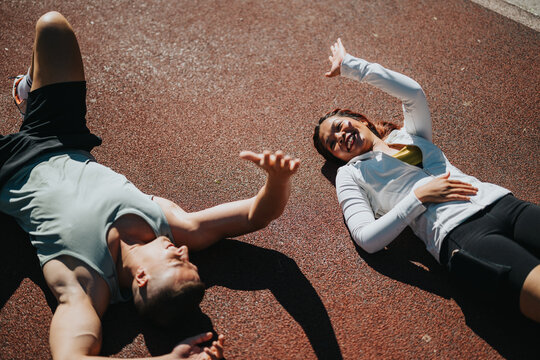 A diverse pair of athletes laughing while lying on a track after exercising, signifying camaraderie, relaxation, and fitness. The image highlights joy and connection through outdoor activities.