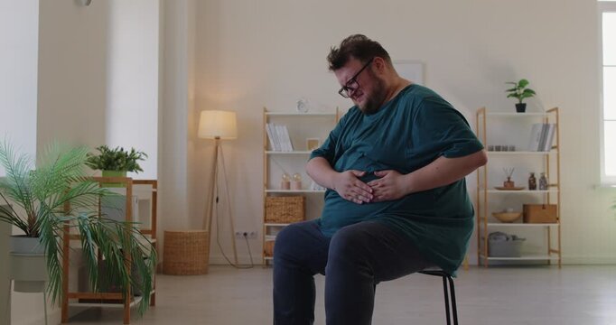 Fat overweight man with glasses sits on a chair at home interior, holding hands on his belly suffering from stomachache. Obese plump male person in casual with abdomen pain in living room.