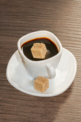 Cup of black coffee and a sugar dropping into it on the surface, served in a white cup on a saucer filled with a brown sugar cube over a wooden table.