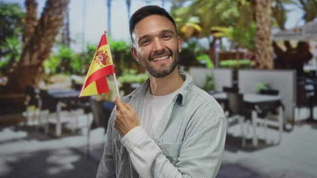 Man holding spain flag smiling outdoors on terrace restaurant surrounded by tables palm trees vibrant background celebrating national pride