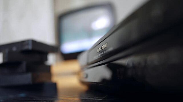 Blurred image of a DVD player, pile of tapes and old TV screen. Close up. Hand presses the buttons.