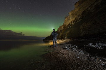 Fototapeta premium A lone adventurer with a headlamp watches the stunning northern lights on a rocky beach by the cliffs of Paldiski, Estonia.