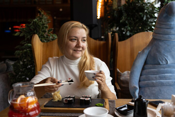 The blonde woman with long blond hair sits at a restaurant table, using chopsticks to eat sushi. A cup and a drink are on the table.