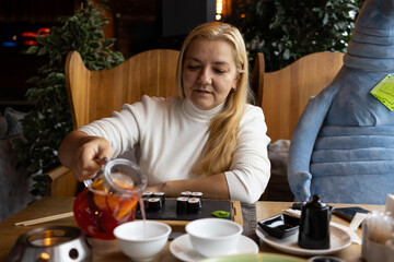 The blonde woman with long blond hair sits at a restaurant table, using chopsticks to eat sushi. A cup and a drink are on the table.