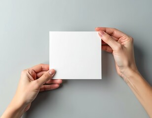 Fototapeta premium Female hands hold a blank square card on gray background. Person presenting a white card with copy space for design or text. Woman showcasing empty card for branding or advertising.