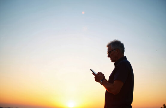 Mature man silhouette texts on smartphone at sunset. Senior uses mobile device for communication on dusk sky background. Person holds cell. Digital lifestyle and connection.