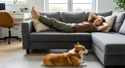 A relaxed person rests on a comfortable gray sofa in a bright living room, with their Corgi dog lying on the floor.