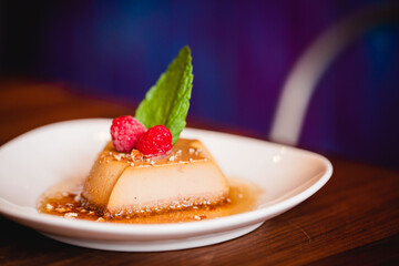 Flan with raspberries and fresh mint on a white plate against a bright and colorful restaurant 