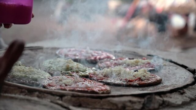 Hands of a mature woman preparing corn gorditas with cheese on a comal in an outdoor kitchen, smoke rising from the comal, Mexican food