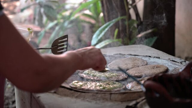 Hands of a mature woman preparing corn gorditas with green sauce on a comal in an outdoor kitchen, smoke rising, Mexican food