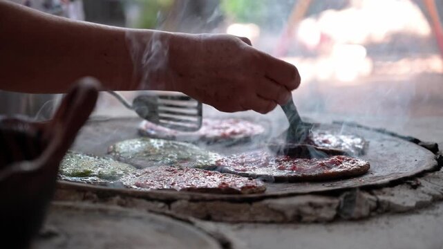 Mexican food gorditas, Hands of a mature woman spreading red sauce on corn gorditas on a clay comal in an outdoor kitchen, smoke rising