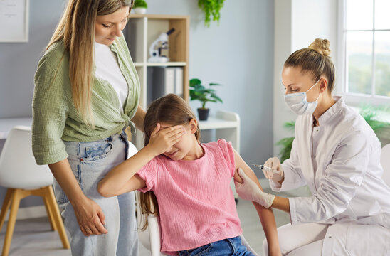 Vaccination injection for scared child by nurse, clinic. A masked nurse administers a shot as the pediatric patient covers her eyes, with mother supporting. Clear message of pediatric safety.