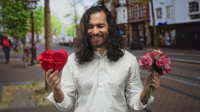 Man holds red heart box and pink bouquet in hands while puffing cheeks on a busy city street; awkward romance.