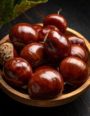Obraz premium Close-up of shiny, round chestnuts in a rustic wooden bowl, displayed on a dark, wooden surface, with a bit of leaf visible