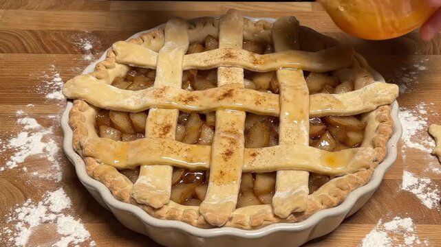 a hand using a pastry brush to apply egg wash or glaze over a lattice crust of an apple pie before baking, showing the final stage of preparation of a delicious sweet homemade dessert.