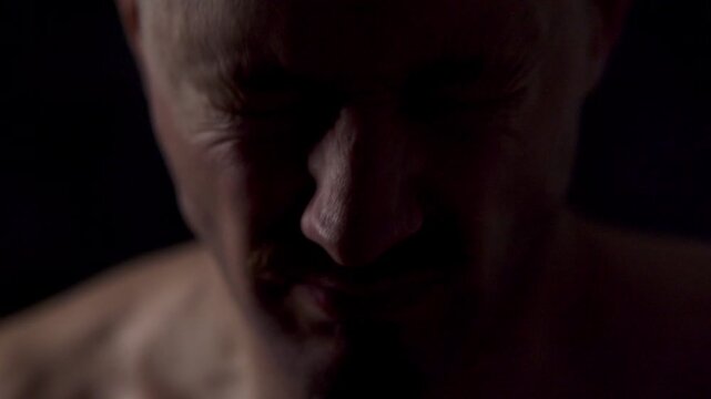 A close-up portrait of an athletic man against a dark background. He stares at the camera intently and slaps his face with his palms to energize himself and regain his composure.