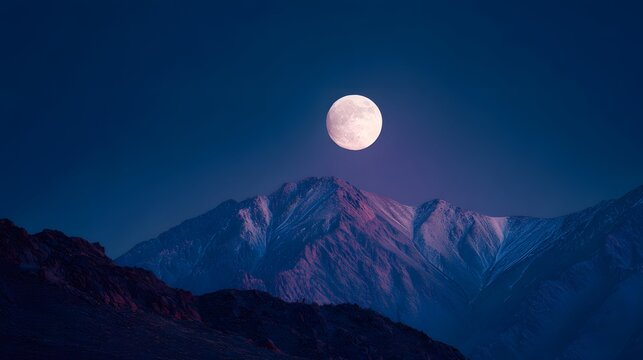 Full moon illuminates rugged snow-capped mountains in a deep blue sky.