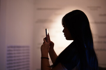 Silhouette of a black-haired young girl holding a smartphone in her hand, looking forward into the room.