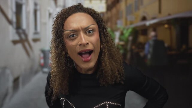 Hispanic transgender woman with curly hair touches cheeks with open mouth on narrow street; surprise.