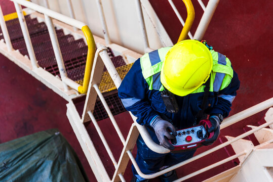 Wearing a yellow hard hat and high-visibility jacket, a seafarer operates a remote control for a provision crane on a merchant ship's deck.