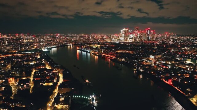 England, London: Canary Wharf skyscrapers reflecting on River Thames at night, aerial view of London urban cityscape with colorful illuminated buildings under dark cloudy sky. Drone flight footage